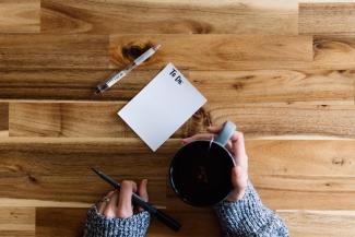 a person writing on a piece of paper next to a cup of coffee by Kelly Sikkema courtesy of Unsplash.